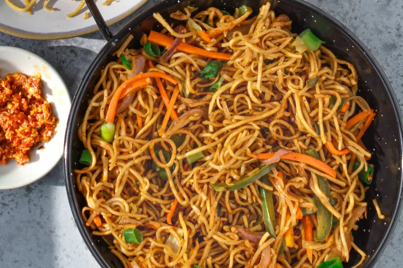 Wok of vegetable Hakka noodles with carrots, bell peppers and scallions, served with a side of noodles and chili garlic sauce, at Hakka Express in Austin