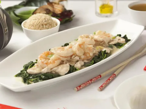 Platter of shrimp and sliced chicken over leafy greens at Bo Lings in Overland Park, served with a striped teapot, a bowl of rice, a cup of tea, and red Lunar New Year decorations on a white table