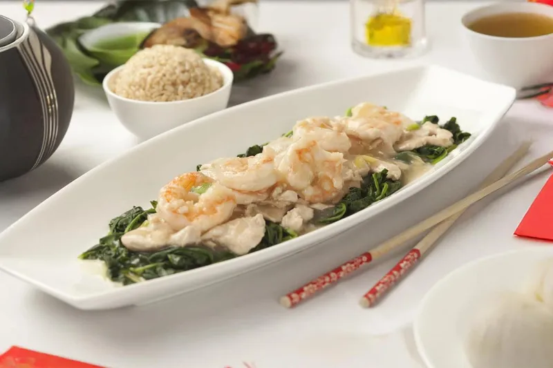 Platter of shrimp and sliced chicken over leafy greens at Bo Lings in Overland Park, served with a striped teapot, a bowl of rice, a cup of tea, and red Lunar New Year decorations on a white table